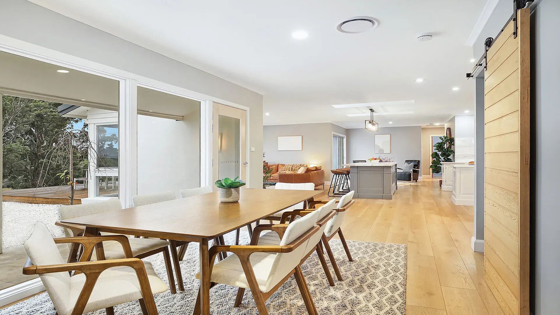 A spacious dining area with a long wooden table and chairs, a kitchen in the background, and a sliding door leading to an outdoor area.