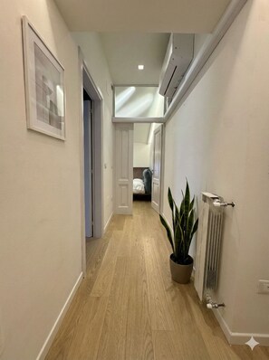 Un pasillo estrecho con suelo de madera, una planta en maceta, un radiador y un cuadro en la pared.