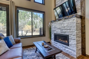 A living room with a stone fireplace, a sofa with cushions, a coffee table, and a large window with a view of trees. - Luxury Black Hills Chalet with Hot Tub Near Mount Rushmore, Keystone SD (Keystone)