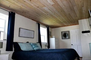 A bedroom with a wooden ceiling, a bed with a dark blue bedspread, a white radiator, and a door. - Charming Dog-Friendly Beach House with Fire Pit & Jetted Tub Near Carters Beach in Hancock, Maine (Hancock)