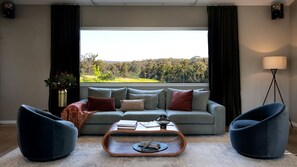 A living room with a grey sofa, two blue armchairs, a coffee table, and a view of a green landscape through a large window.