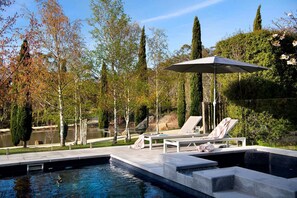 A swimming pool with two lounge chairs and an umbrella, surrounded by trees and a view of a body of water.
