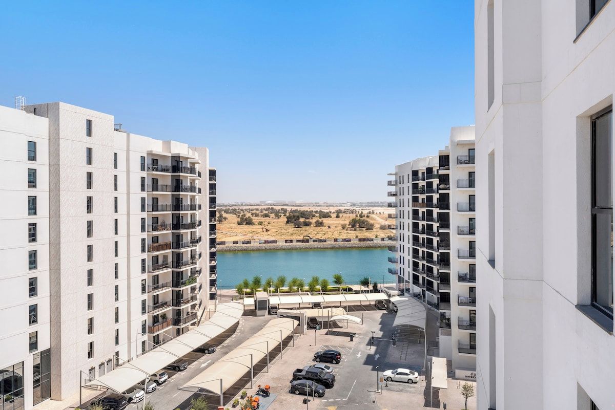 A high-rise residential building with multiple balconies, a parking area, and a view of a body of water and distant landscape.