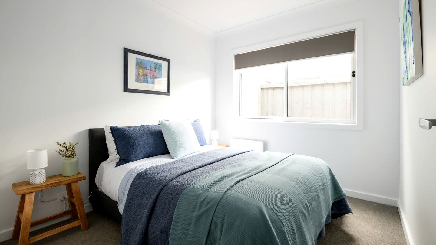 A bedroom with a bed, a bedside table with a lamp, a framed picture on the wall, and a window with blinds.