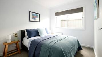 A bedroom with a bed, a bedside table with a lamp, a framed picture on the wall, and a window with blinds.
