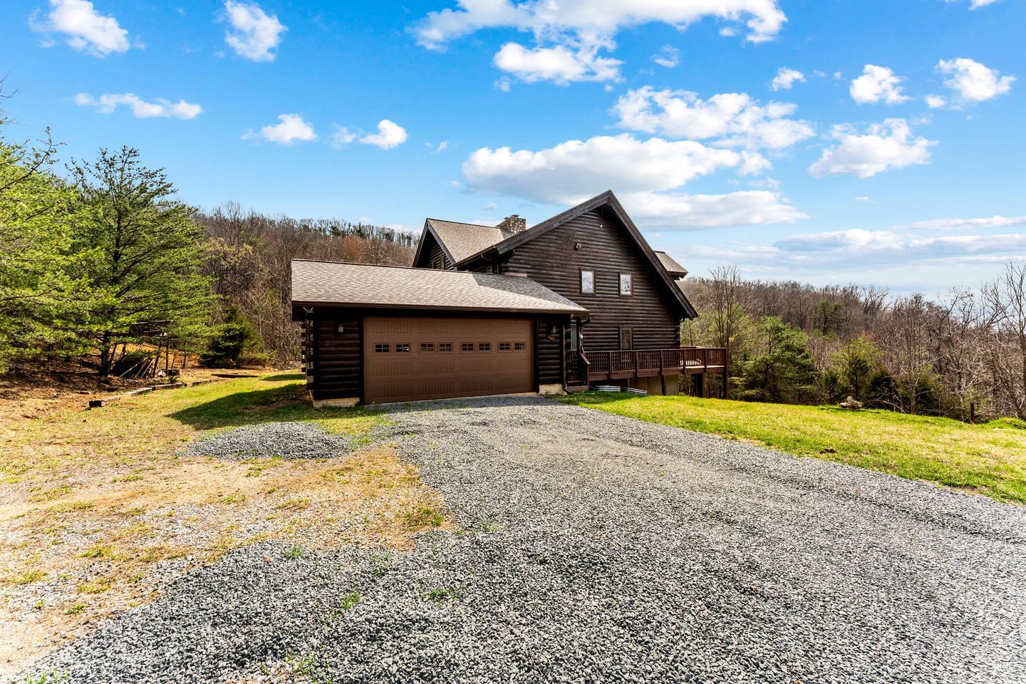 A log cabin with a garage, surrounded by a gravel driveway and grassy areas, with trees and a clear sky in the background.
