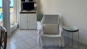 Um quarto com ventilador de teto, uma poltrona branca, uma mesinha, um armário com TV e vista por uma porta de vidro.