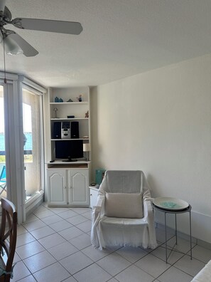A room with a ceiling fan, a white armchair, a small table, a cabinet with a TV, and a view through a glass door. - Apartment with a view of the pond and mountains, featuring a pool (Le Barcarès)