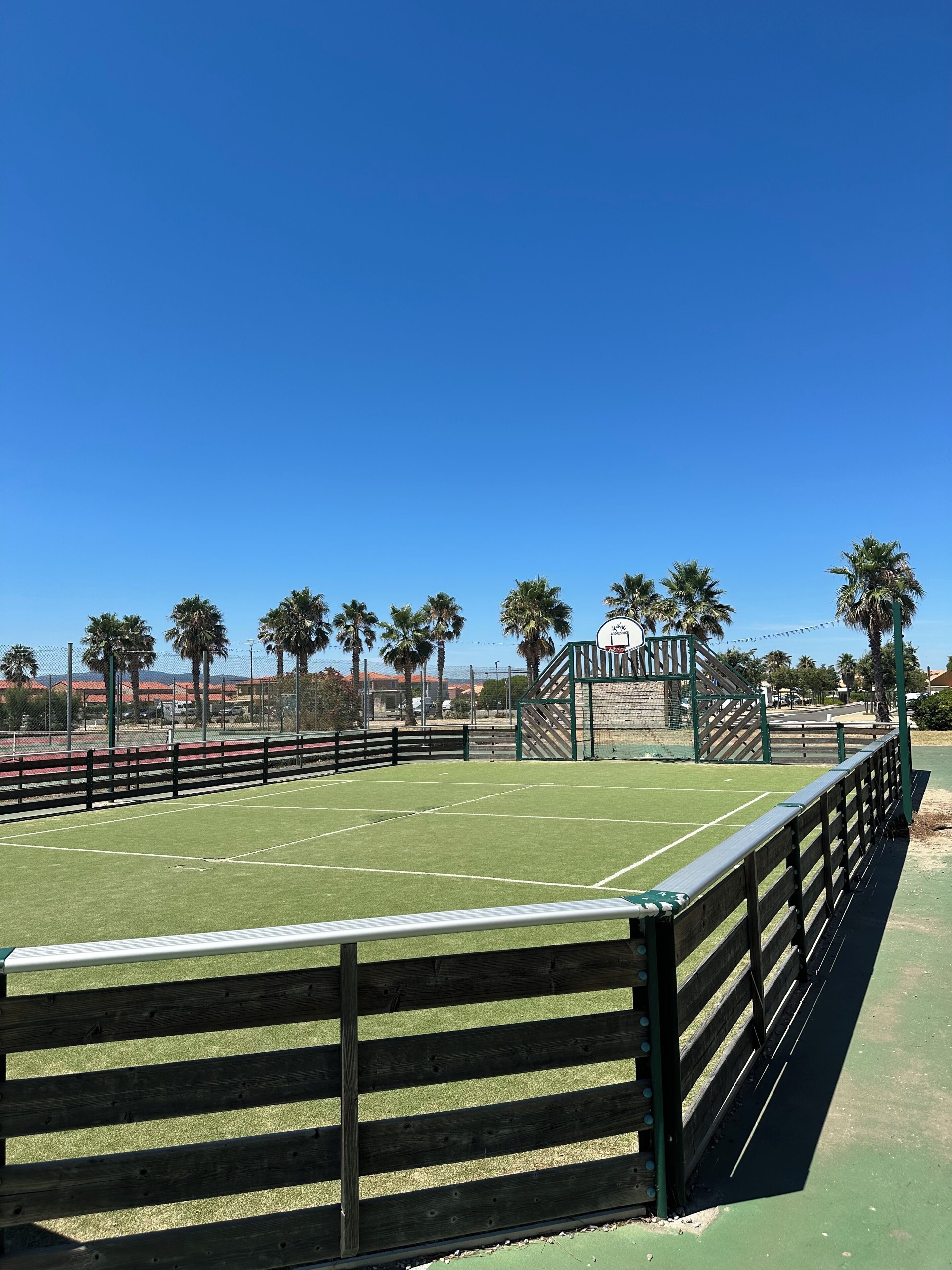 A tennis court with a wooden fence, palm trees, and a clear blue sky.