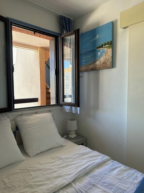 A bedroom with a bed, a nightstand with a lamp, a window with curtains, and a painting on the wall. - Apartment with a view of the pond and mountains, featuring a pool (Le Barcarès)