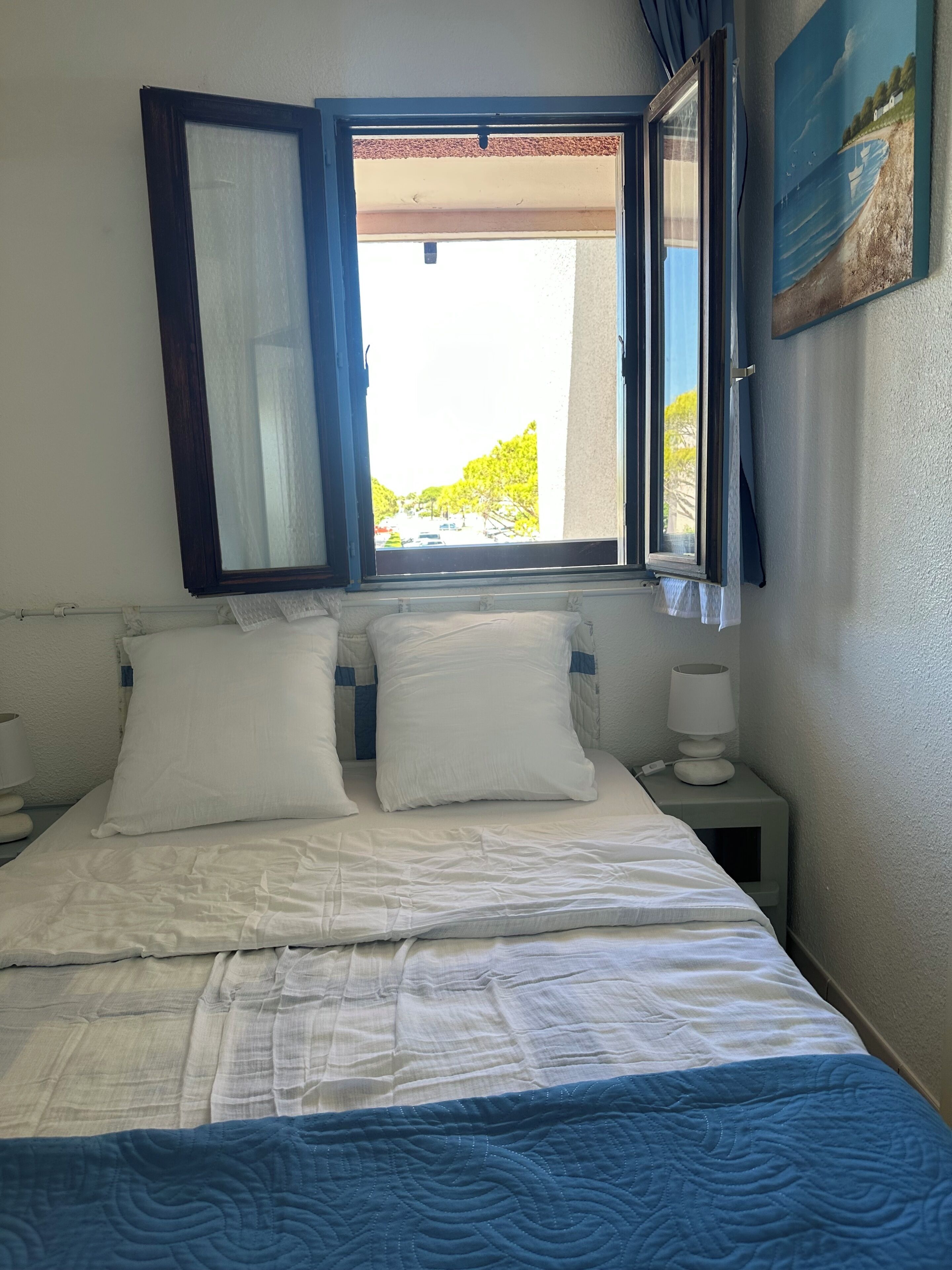 Un lit avec une literie blanche, une couverture bleue et une fenêtre donnant sur des arbres et un immeuble.