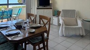 Uma sala de jantar com uma mesa posta para quatro pessoas, uma televisão em um suporte, um ventilador de teto e vista para o mar através de uma porta de vidro.