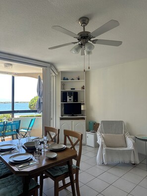 A dining area with a table set for four, a television on a stand, a ceiling fan, and a view of the sea through a glass door. - Apartment with a view of the pond and mountains, featuring a pool (Le Barcarès)
