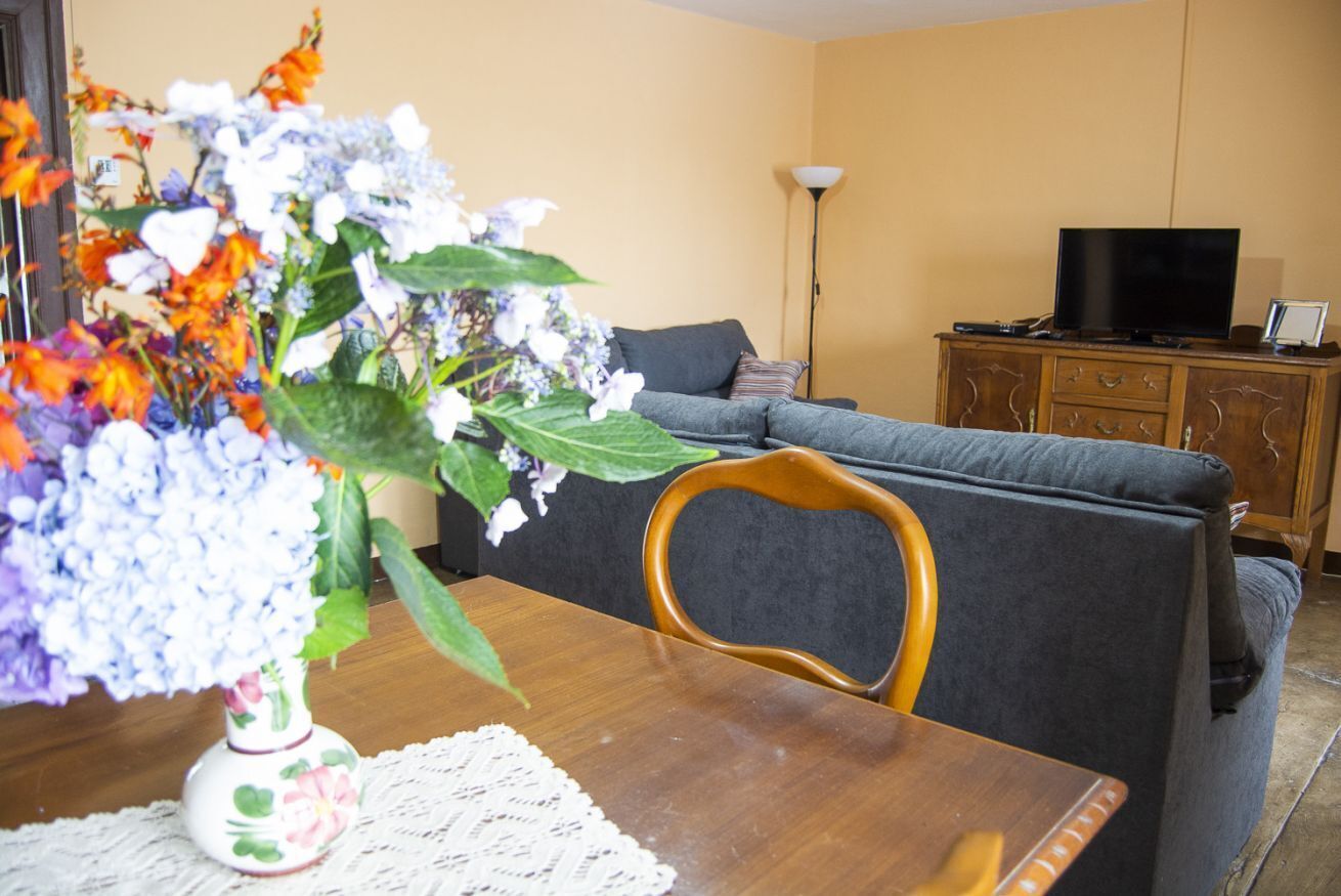 A living room with a sofa, a television on a wooden cabinet, and a table with a vase of flowers.