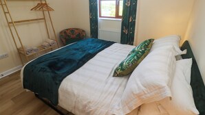 Una cama con un edredón azul oscuro y almohadas blancas, un perchero de madera con toallas dobladas, un sillón estampado y una ventana con cortinas verdes.