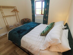 Una cama con un edredón azul oscuro y almohadas blancas, un perchero de madera con toallas dobladas, un sillón estampado y una ventana con cortinas verdes.