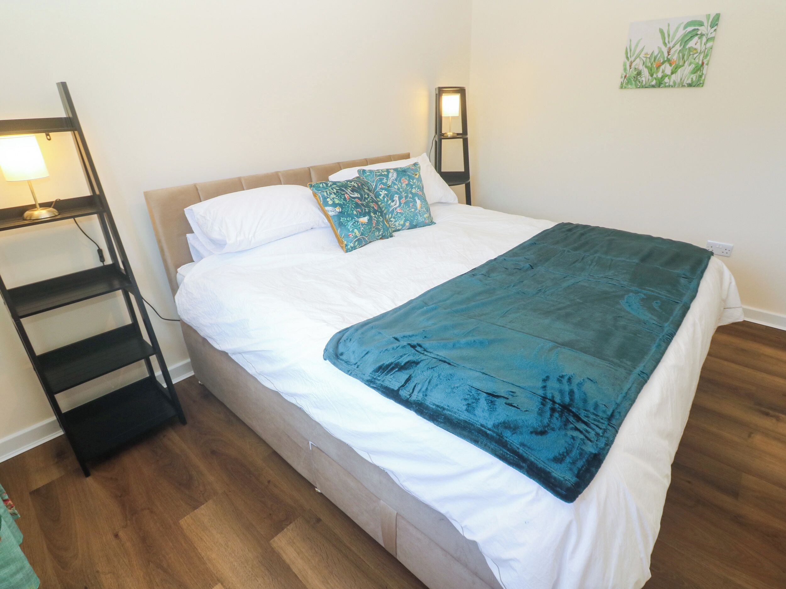 Una cama bien hecha con edredón blanco y manta verde aguamarina, una lámpara de noche y una mesita de noche fijada a la pared con una lámpara.