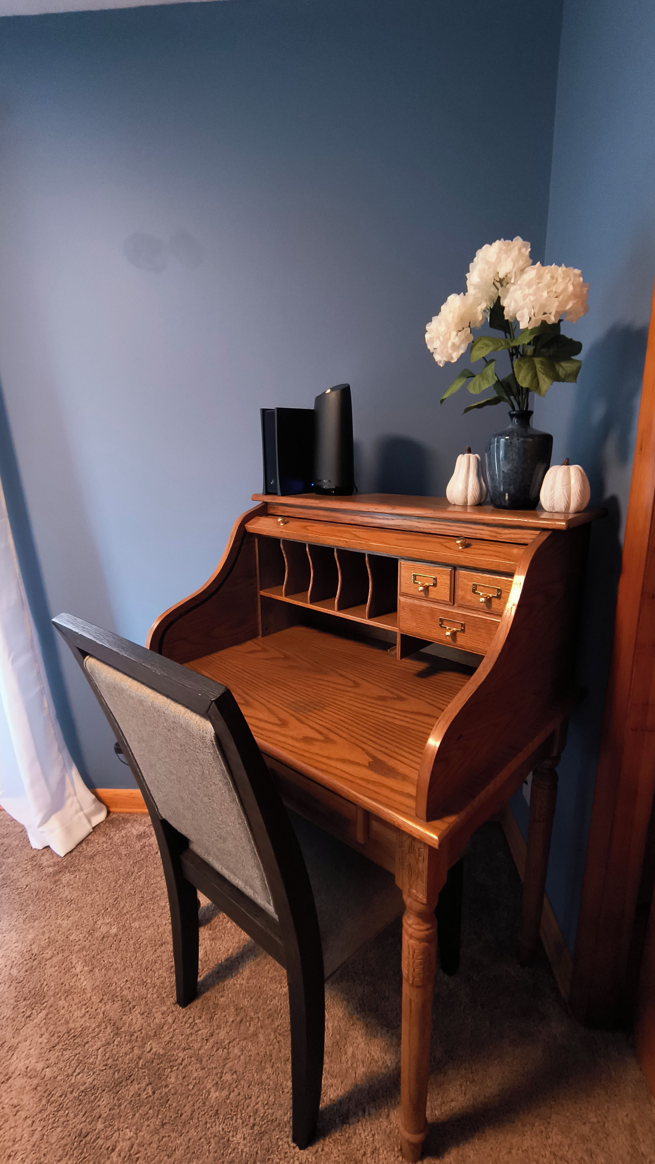 Een houten bureau met een stoel, een vaas met bloemen en een zwart voorwerp op het bureau.