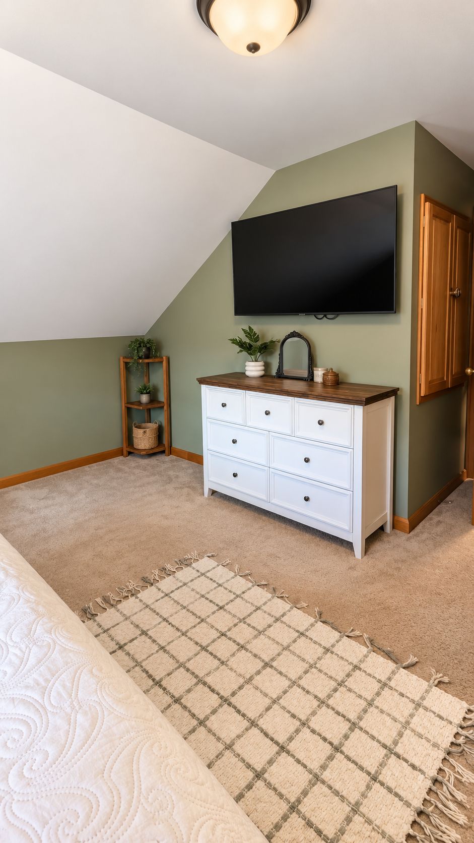 A bedroom with a sloped ceiling, a flat-screen TV mounted on the wall, a white dresser with drawers, a wooden cabinet, a small plant shelf, and a checkered rug.