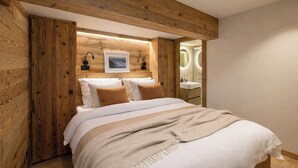 A bedroom with a wooden headboard, a bed with white bedding and brown pillows, a bedside table with a lamp, and a bathroom visible through an open door. - Chalet Montâme - Modern Luxury in the Swiss Alps (Riddes)