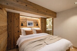 Ein Schlafzimmer mit Holz-Kopfteil, einem Bett mit weißer Bettwäsche und braunen Kissen, einem Nachttisch mit Lampe und einem durchgegangenen Tür angedeuteten Badezimmer.