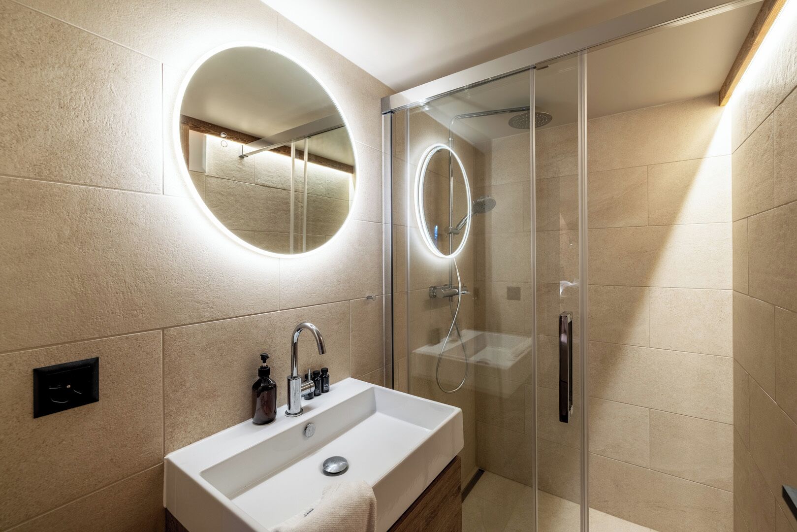 Un baño moderno con un lavamanos grande de color blanco, una ducha con mampara de vidrio y un espejo redondo con iluminación.