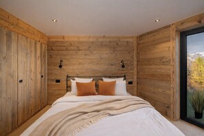 Een slaapkamer met een houten wandpaneel, een bed met witte beddengoed en twee oranje kussens, en een raam met uitzicht op bomen en bergen.