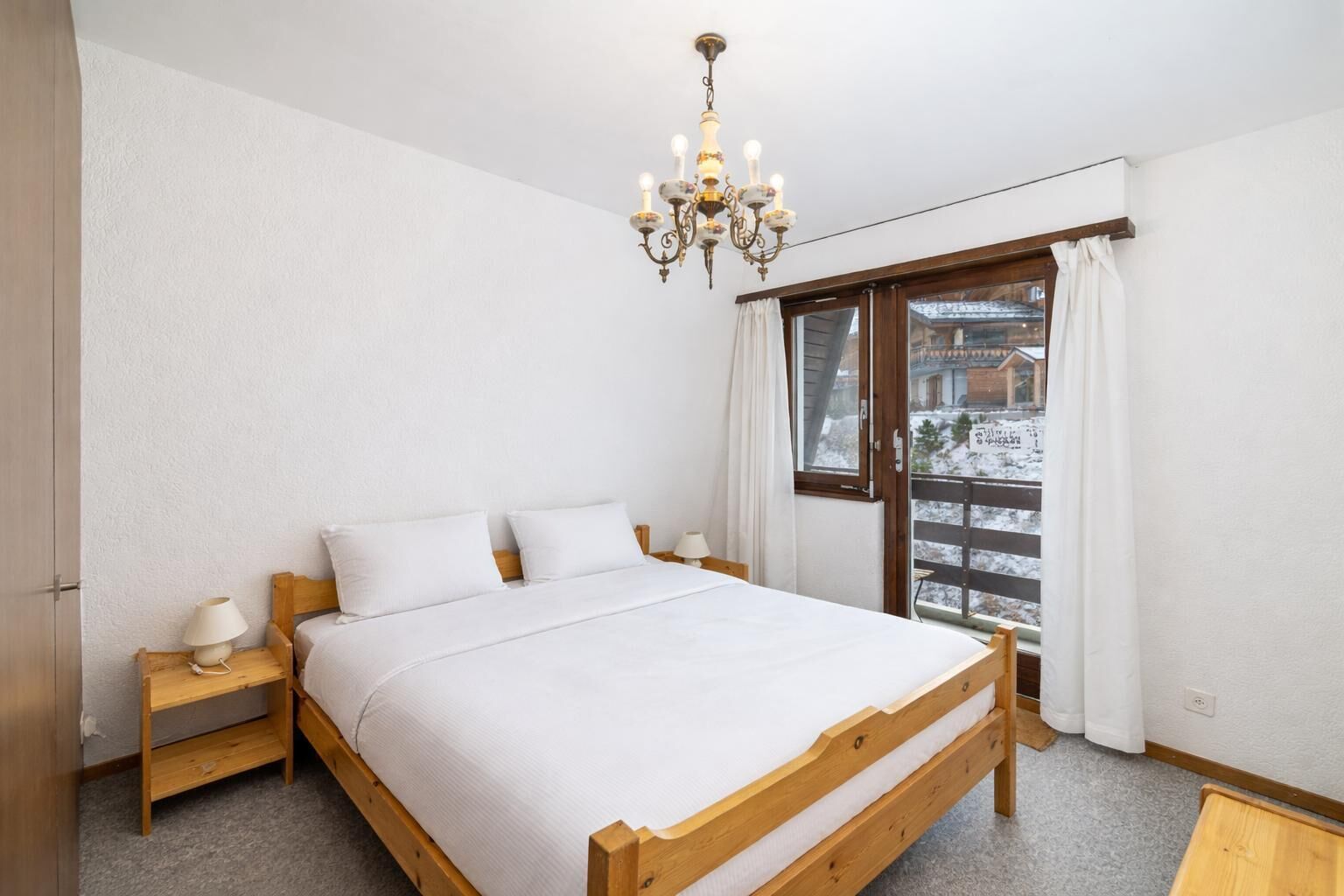 Une chambre à coucher avec un lit en bois, une table de chevet, une lampe, un lustre, une fenêtre avec des rideaux et une vue sur un paysage enneigé.
