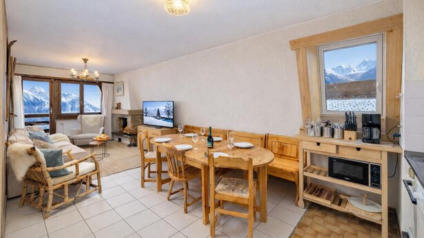 A cozy living room with a fireplace, a dining area with wooden furniture, and a kitchen with a built-in oven and a window with a mountain view. - Apartment Mont-parrain C28 Cosy & Central With Private Parking (Riddes)