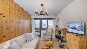 A cozy living room with a sofa, a chair, a small table with a drink, a fireplace, and a television displaying a snowy landscape. - Apartment Mont-parrain C28 Cosy & Central With Private Parking (Riddes)