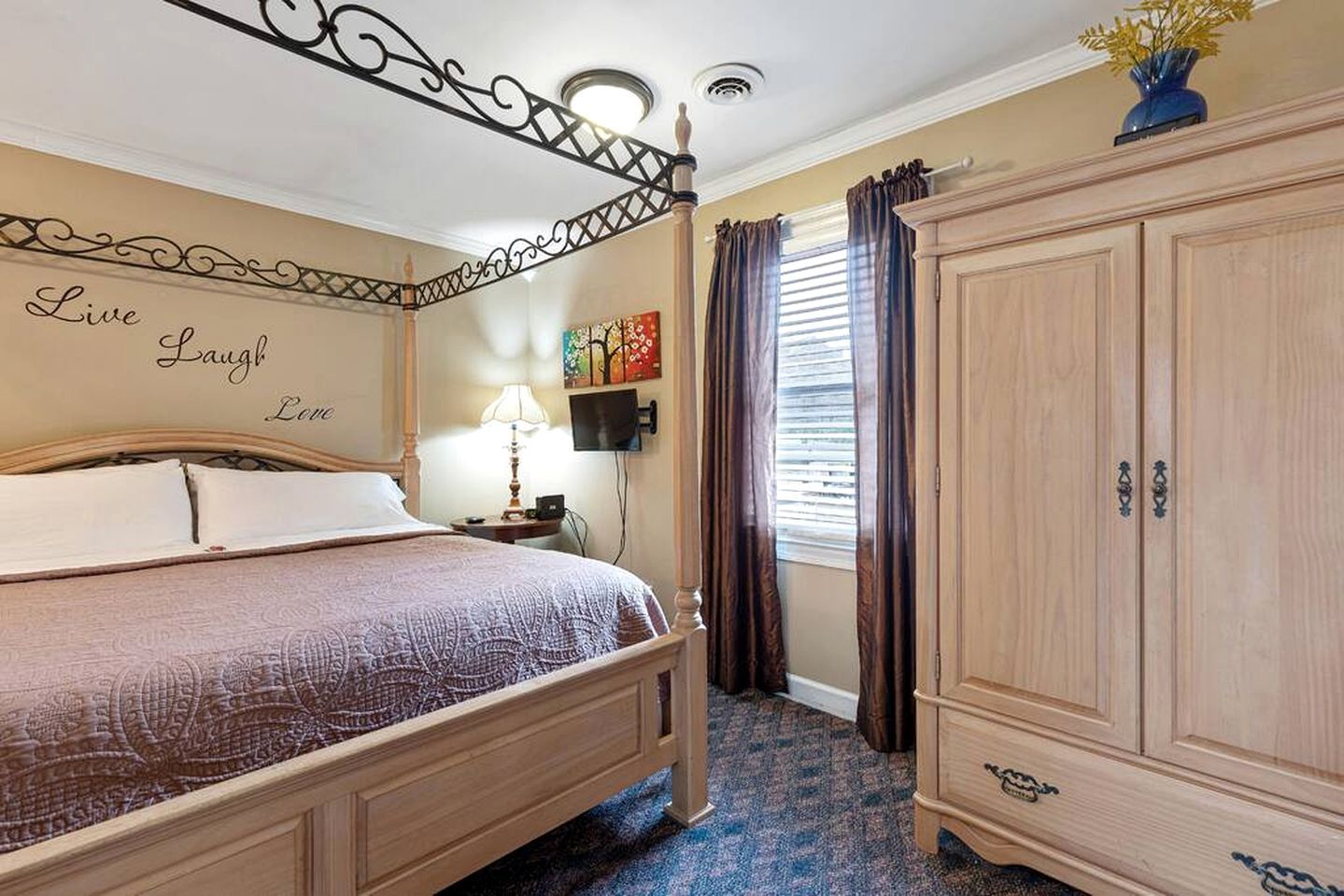 A bedroom with a bed, bedside table, lamp, wardrobe, and a wall with a decorative phrase.