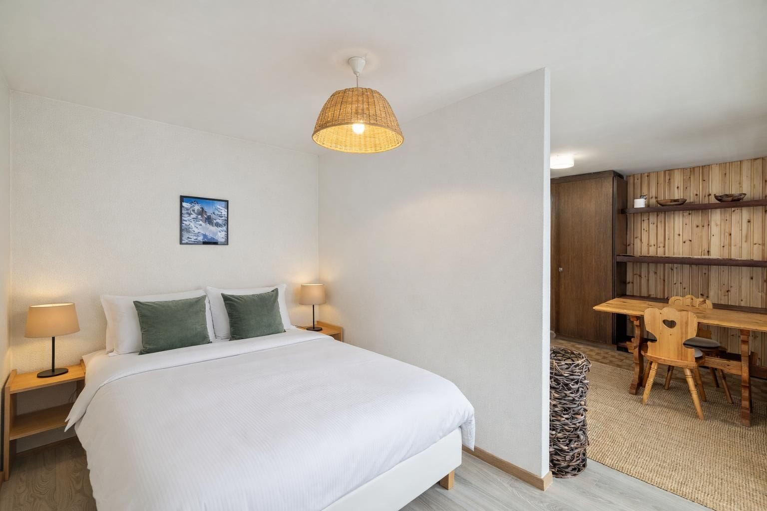 Een slaapkamer met een bed, bedlampen, een hanglamp, een schilderij en een houten eethoek.