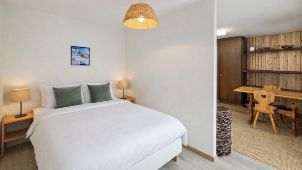 A bedroom with a bed, bedside lamps, a hanging light fixture, a painting, and a wooden dining area. - Apartment Mont-parrain C32 Bright & Central With Balcony (Riddes)