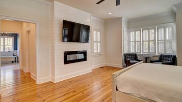 A bedroom with a bed, a television mounted on the wall, a ceiling fan, and a window with shutters.