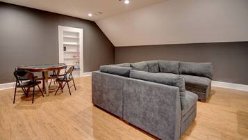 A room with a grey sectional sofa, a round wooden table with chairs, and a white shelving unit against a grey wall.