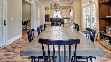 A spacious dining area with a long table and chairs, a chandelier, and a kitchen in the background.