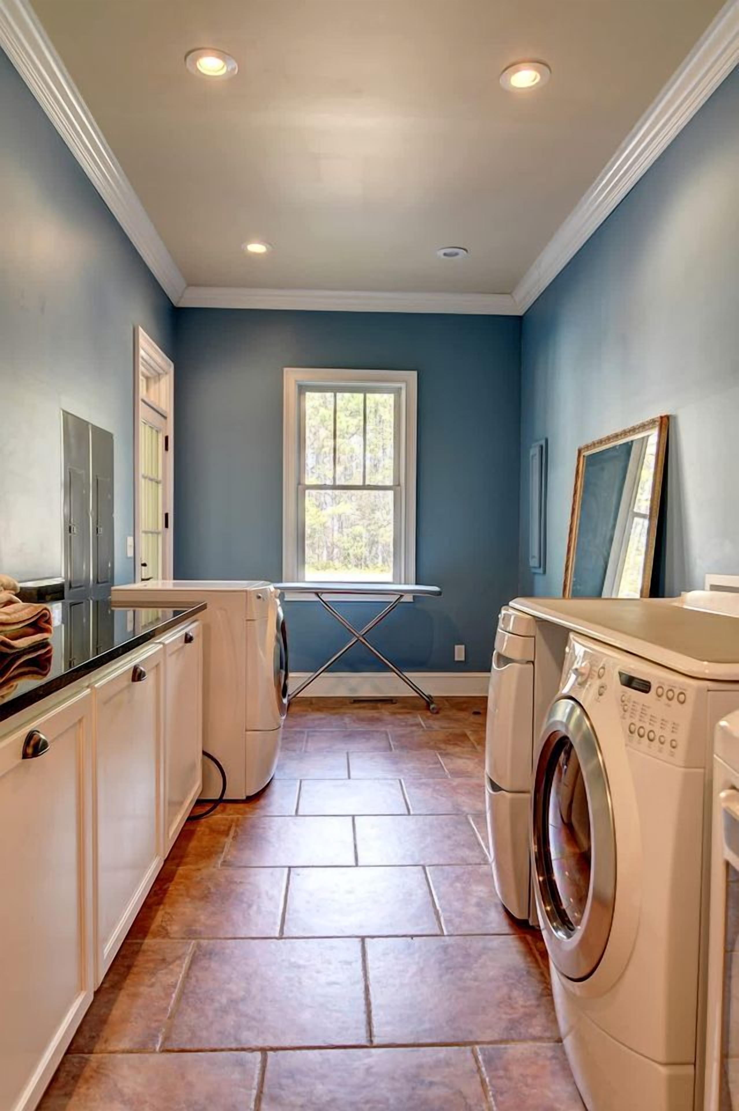 A laundry room with a washing machine, dryer, ironing board, and a window providing natural light.