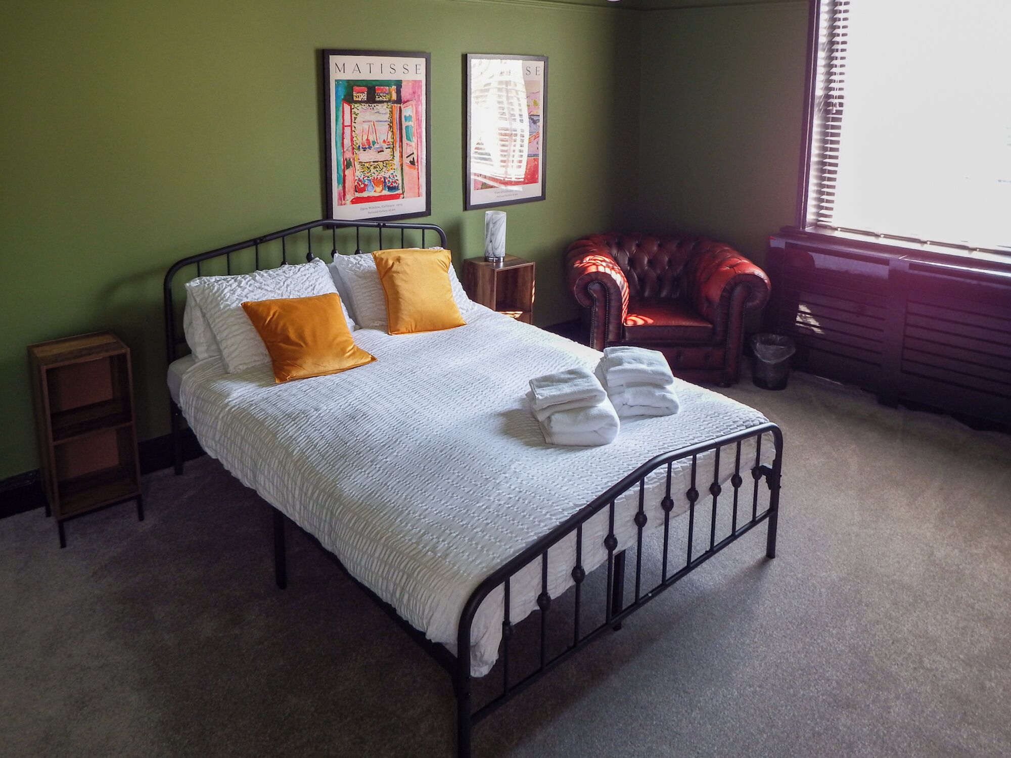 Una cama bien hecha con un edredón blanco y dos almohadas naranjas, un sillón de cuero rojo, y dos cuadros en la pared.