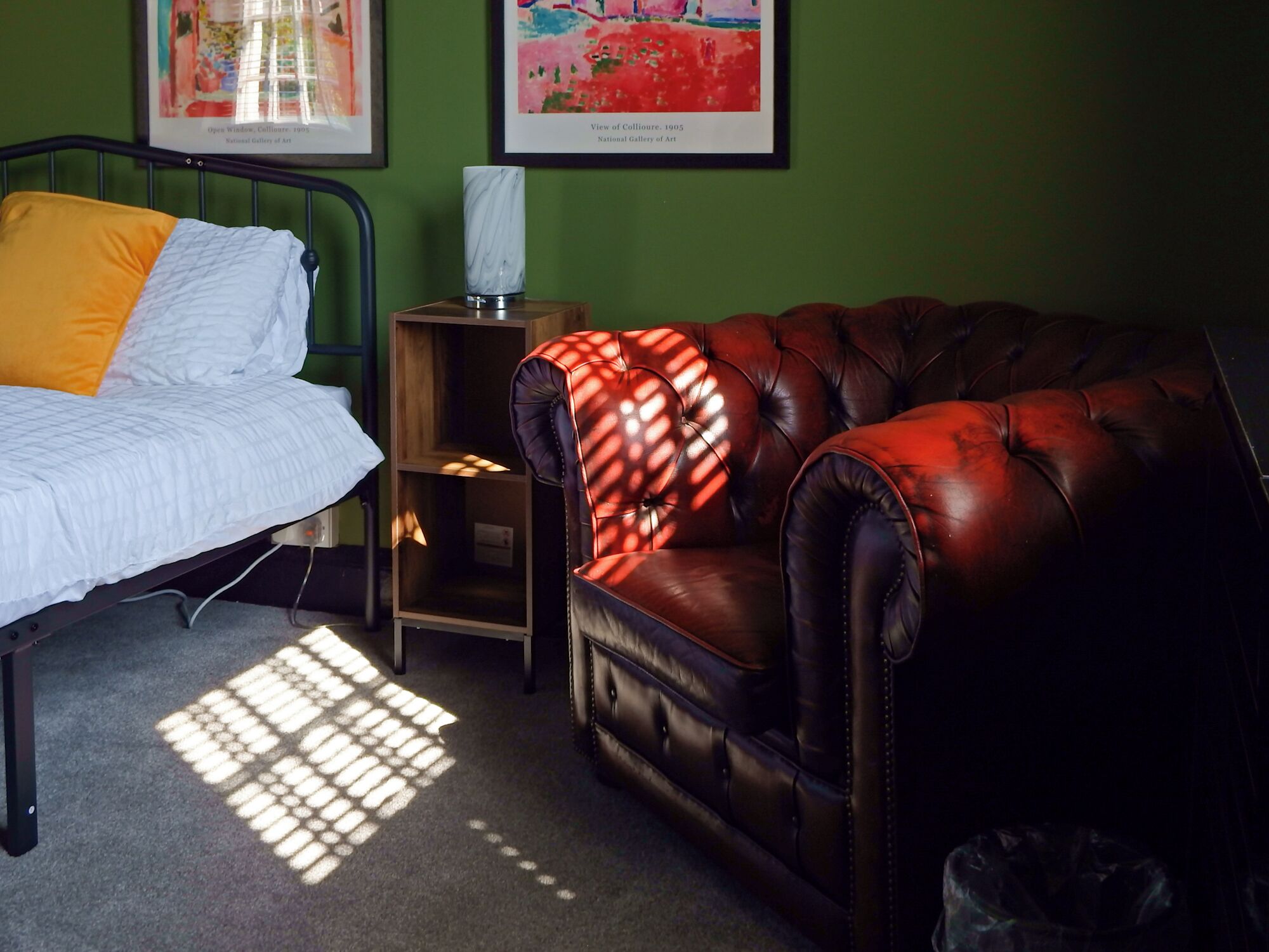 A bedroom with a bed, a leather armchair, a wooden nightstand, and two framed artworks on the wall.