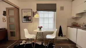 Eine kleine Küche mit weißen Schränken, einem Esstisch mit zwei Stühlen, einem Heizkörper und einem Fenster mit Vorhang.