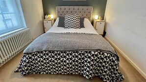Ein Schlafzimmer mit einer gesteppten Kopfteil, einem gemusterten Bettüberwurf, zwei Nachttischlampen, einem gerahmten Bild an der Wand und einer Heizung.