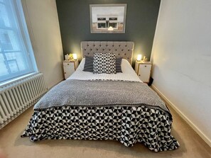 Ein Schlafzimmer mit einer gesteppten Kopfteil, einem gemusterten Bettüberwurf, zwei Nachttischlampen, einem gerahmten Bild an der Wand und einer Heizung.