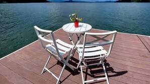 Um deck de madeira com uma mesa redonda e duas cadeiras, com vista para um lago calmo e margens arborizadas.