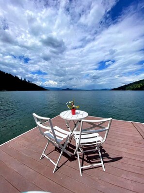 Une terrasse en bois avec une table ronde et deux chaises, donnant sur un lac paisible dont les rives sont bordées d’une forêt.