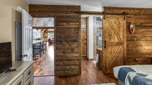 Una habitación con puerta de madera que da a un comedor, cama con colcha estampada y televisor sobre un aparador.