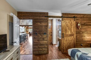 Um quarto com porta de madeira que dá para uma área de refeições, uma cama com colcha estampada e uma televisão sobre um aparador.