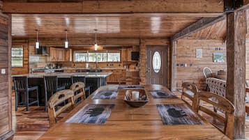 Interior de una cabaña de madera con comedor, cocina y un ventanal grande.