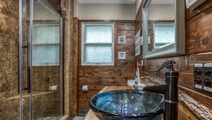 Un baño con lavamanos de vidrio, paredes de panel de madera, cabina de ducha y tocador con espejo.
