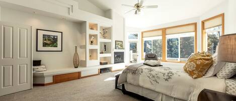 A bedroom with a bed, a seating area with a bench, a fireplace, and large windows.