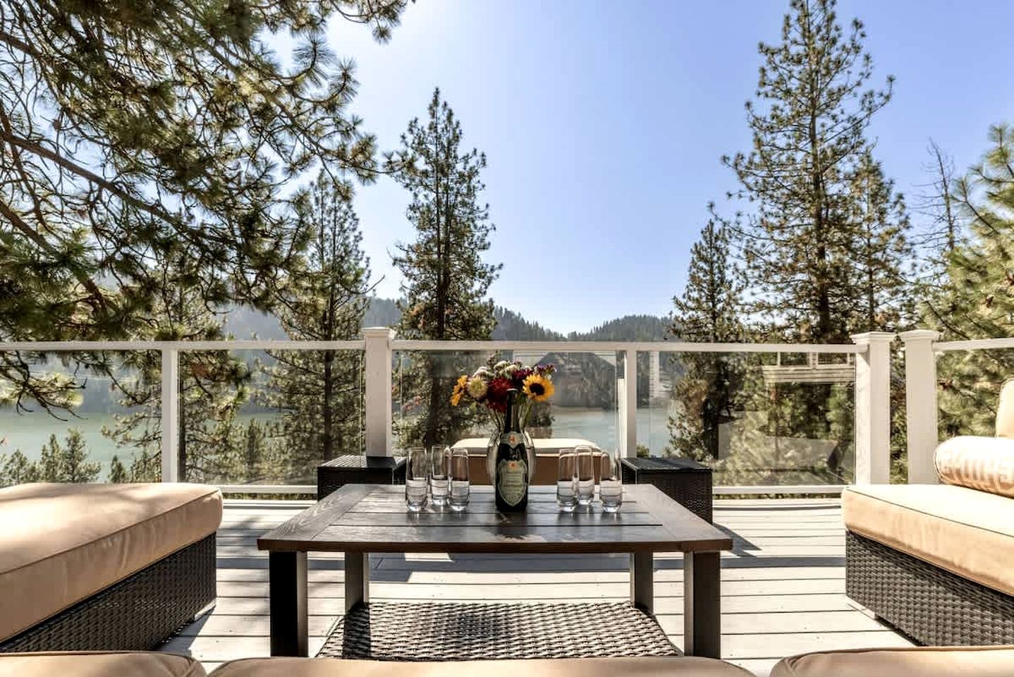 A deck with outdoor furniture, a table with a vase of flowers, and a view of a lake surrounded by trees.